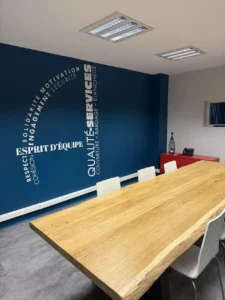 Salle de réunion d’Applitech avec une grande table en bois et un mur bleu mettant en avant les valeurs de qualité, services et esprit d’équipe sous la forme du logo d'Applitech Enveloppe.