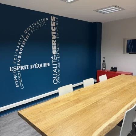 Salle de réunion d’Applitech avec une grande table en bois et un mur bleu mettant en avant les valeurs de qualité, services et esprit d’équipe sous la forme du logo d'Applitech Enveloppe.