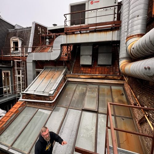 Vue en surplomb d'une verrière de toiture ancienne et dégradée à Lille. On aperçoit des vitrages encrassés et jaunis par le temps, une structure métallique oxydée et un technicien d'Applitech Enveloppe inspectant la zone à proximité d'installations techniques industrielles (CVC).